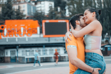 Muchacho y joven chica besándose abrazados en una plaza de la ciudad junto a los arboles y la...