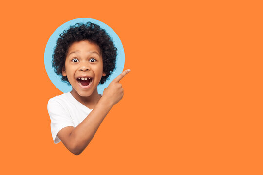 Wow look advertise here! Portrait of amazed little boy with curly hair pointing to empty place on background showing copy space for promotional ad. indoor isolated in a round hole on orange background