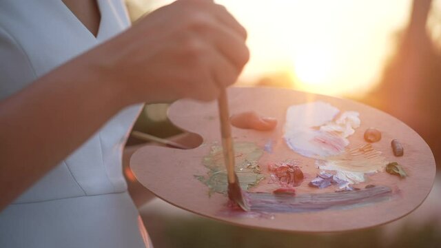 Unrecognizable Caucasian woman mixing colors with brush in slow motion on palette in sunrays. Slim female painter enjoying painting outdoors at summer sunset. Creativity and art concept