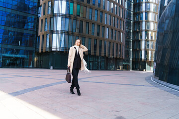 Fototapeta na wymiar A successful Caucasian woman in a coat and suit and glasses stands in front of an office building with a bag for documents. Business woman concept. Business district of the city