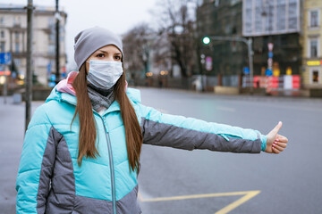 a young woman of Caucasian ethnicity in a warm jacket and hat stands on the street in a mask, stops the car with a hand gesture on the road on a cold cloudy winter day