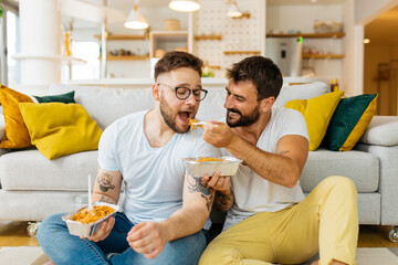 Attractive happy married hipster couple sitting on the floor in the living room and eating pasta for dinner.