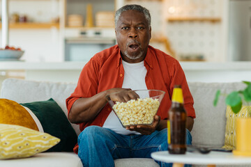 African pensioner sitting at his cozy home, watching television and eating popcorn. He is enjoying the afternoon.