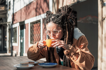 young white hispanic latin girl with glasses and dreadlocks sitting at a table outside a coffee shop drinking a cup of coffee.