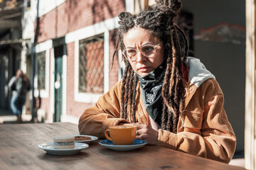 young white hispanic latin girl with glasses and dreadlocks enjoys sitting at a table in a street cafe with a cup of coffee and cookies.