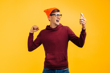 stylish young guy in an orange hat with glasses and a red sweater guy rejoices at a message on the phone