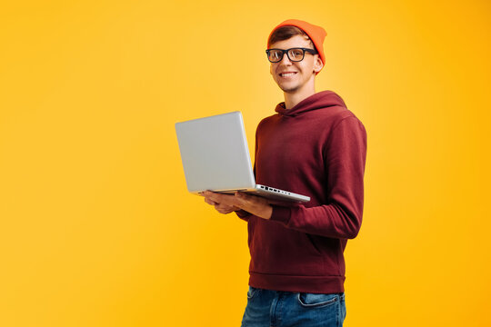 young handsome guy with typing on a laptop, in an orange hat with glasses and a red sweater, on a yellow background