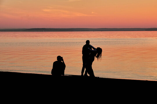 Silhouettes of guy who takes photos on cell phone of couple guy and girl hugging against the background of sunset over the sea