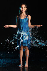 Bright slender beautiful and playful dark-haired teenage girl 12 years old in a blue dress barefoot in a black aqua studio. Emotions in the pouring night rain.