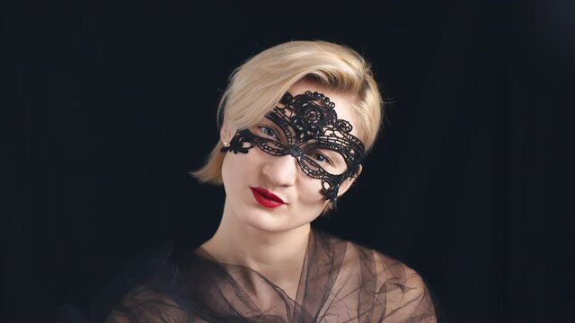 Sexy blond woman with a lace Venetian black mask and red lipstick smiling to the camera. Shoulders covered in a black veil