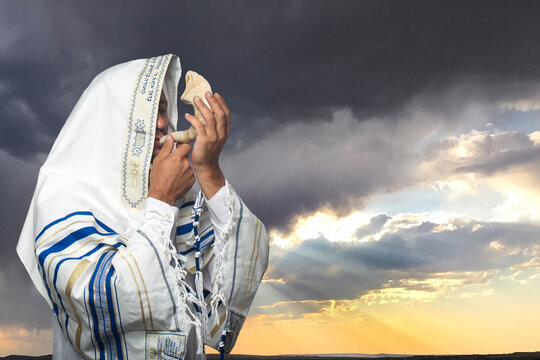 Jewish man wrapped in tallit, prayer shawl with the inscription "Baruch Atah Adonai" written on it in Hebrew, holding the Shofar, ram's horn of Rosh Hashanah (New Year), Yom Kippur on dramatic sunset