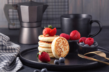 Cottage cheese pancakes with fresh with fresh strawberry and blueberries on dark ceramic plate. Cup of coffee and coffee maker