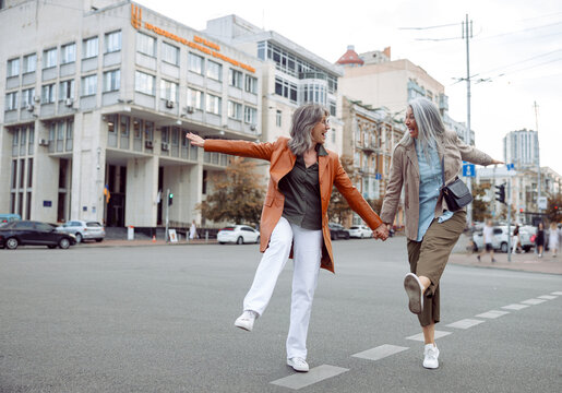 Couple of playful senior women have fun walking along contemporary city street on autumn day. Friends spend time together