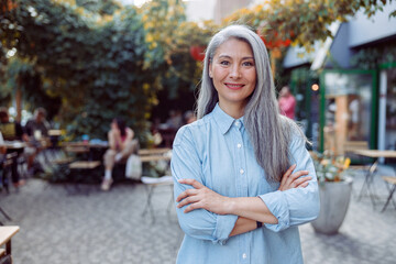 Pretty silver haired senior Asian woman with crossed arms stands against outdoors cafe terrace on...