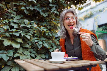 Cheerful hoary haired senior woman with spectacles eats strawberry dessert sitting at table on...