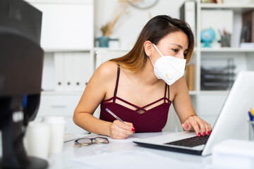 ..Portrait of female worker in disposable face mask engaged in business activities at workplace in...