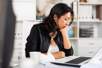 Portrait of frustrated female entrepreneur sitting at office desk with papers and laptop