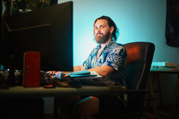 Wide shot of Bearded man use computer against teal light sitting on chair looking at camera