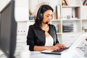 Focused colombian woman call center operator receiving calls and using laptop in office