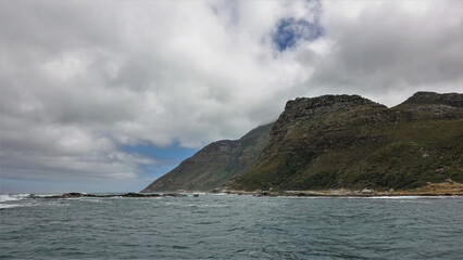 A picturesque cliff on the Atlantic Ocean coast. There is scant vegetation on the steep rocky slopes. The waves are foaming on the stones. Fluffy cumulus clouds float in the blue sky. Cape Town. 