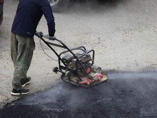 Obraz premium A worker rams new asphalt with a manual motor asphalt rammer, asphalting by the pit method, local road repair