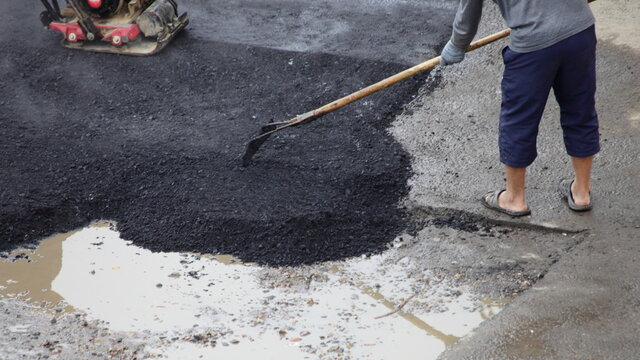 Road worker levels new asphalt in a puddle with a rake on asphalt rammer background, incorrect asphalting by the pit method, local road repairs