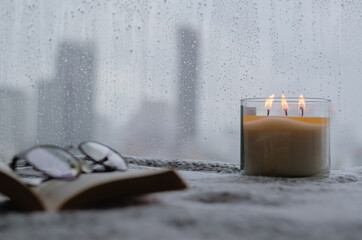 Burning aroma candle puts near by window that have rain drop in monsoon season with blurred city...