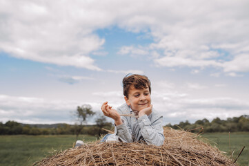 Naklejka na ściany i meble cute schoolboy boy is lying on large haystack in field. happy childhood in the country. games in field. autumn landscape. selective focus