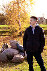 young man in black jacket is walking in an autumn park, standing against background of large stones and autumn birches. Golden Autumn