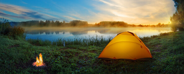 Orange tent with bonfire on bank of misty river at sunset