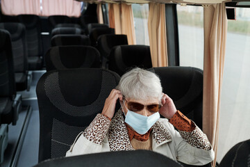 Senior woman in sunglasses sitting in empty bus and putting mask before ride during coronavirus pandemic
