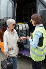 Smiling senior Caucasian woman with suitcase giving ticket to bus conductor in green vest