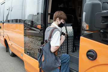 Teenage girl with short hair wearing facial mask holding satchel while getting on contemporary bus