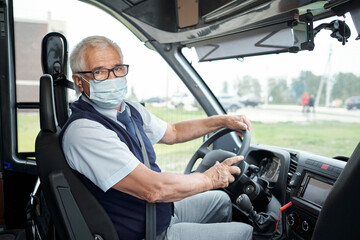 Obraz premium Portrait of senior white-haired bus driver in eyeglasses and facial mask fasten seat belt sitting at wheel