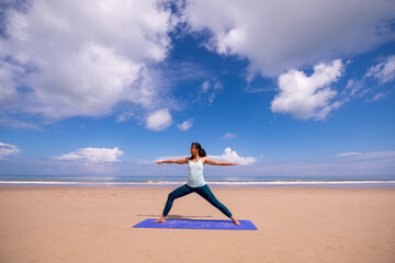 people, fitness, sport and healthy lifestyle concept - young asian woman making warrior yoga poses on tropical beach with blue sky background