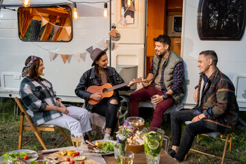 Group of happy young friends singing songs by house on wheels while girl playing guitar