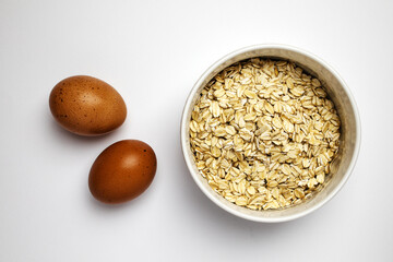 Oats in a bowl and two raw brown eggs aside, top view on white background