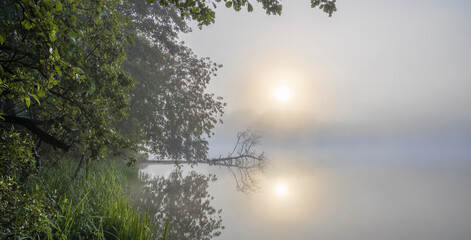 Sunrise over the foggy lake with the reflection of sun. Mist on the water, forest silhouettes and the rays of the rising sun. Beautiful morning landscape with sunrise over river.