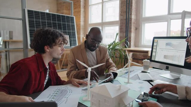 Group of diverse engineers sitting at office table, using digital tablet and working with paper plans while discussing project of alternative energy