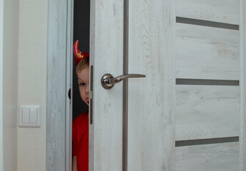 A beautiful little devil with horns. Kid. The concept of Halloween, holiday and childhood. A funny child in a carnival costume looks out from behind the door of the house. Selective focu.