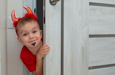 A beautiful little devil with horns. Kid. The concept of Halloween, holiday and childhood. A funny child in a carnival costume looks out from behind the door of the house. Selective focu.