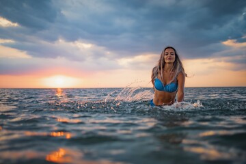 A girl with blond hair in a blue swimsuit splashes to the sides while sitting in an estuary on a sunset background