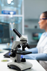 Selectiv focus on medical microscope standing on table in biochemistry hospital laboratory. Specialist microbiologist woman doctor working at biology experiment typing healthcare expertise
