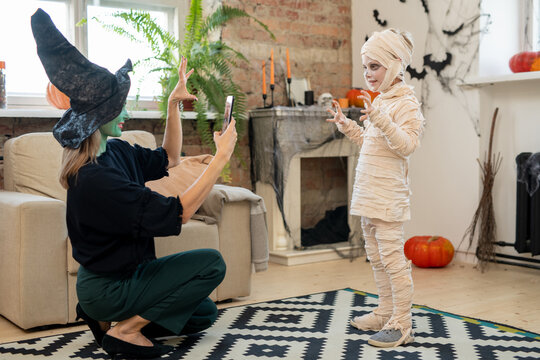 Female in witch costume taking photo of her little son in zombie attire in living-room