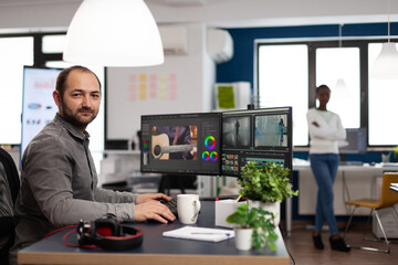 Creative videographer artist man looking at camera smiling working in creativity multimedia startup office using editing post production software on professional pc. Processing video film montage.