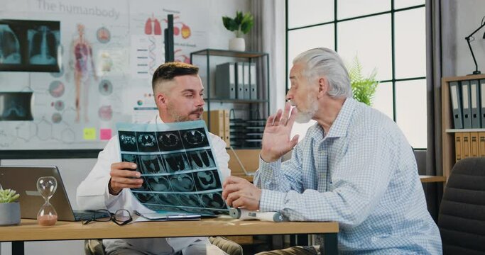 Medicine concept where handsome successful qualified bearded doctor explaining results of x-ray scan for attantive respected senior bearded patient during appointment in clinic