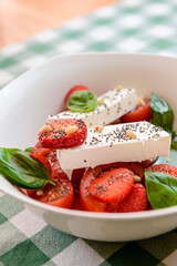 Caprese salad with strawberry, mozzarells, basil and balsamic sauce served in a white bowl over green plaid background.