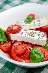 Caprese salad with strawberry, mozzarells, basil and balsamic sauce served in a white bowl over green plaid background.