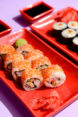 Japanese sushi rolls served on a red plate with pickled ginger and wasabi sauce over colorful purple background.
