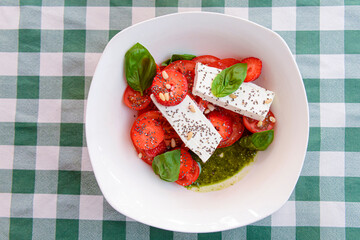 Caprese salad with strawberry, mozzarells, basil and balsamic sauce served in a white bowl over green plaid background.
